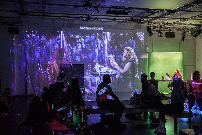 A darkened room with people seated on benches watching a large projection on a translucent screen. The projection shows two persons in conversation, one holding a drink, with the subtitle "Do you want some?" displayed above them. The scene has a surreal, colorful overlay, blending purple and blue tones. In the background, a person with curly hair sits at a table with a bottle, lit by green light, creating an immersive, theatrical atmosphere.