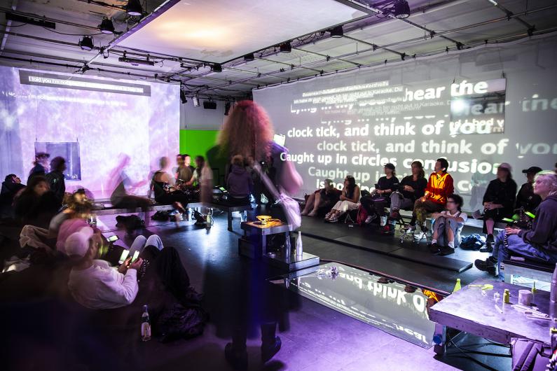 A dimly lit performance space filled with people seated on benches or lounging on the floor. The background features a projection of large, blurry white text against a purple-tinted screen. The atmosphere is casual and immersive, with soft lighting and a reflective floor adding to the ambiance.