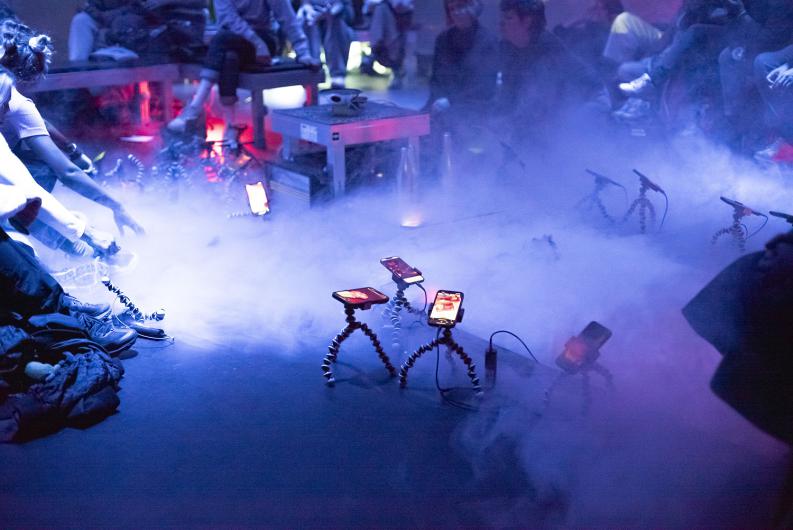 Eine Nahaufnahme einer Performance-Installation mit Smartphones, die auf flexiblen Stativhaltern montiert sind und in einer nebligen, blau beleuchteten Umgebung leuchten. Publikumsgäste sitzen auf Bänken und interagieren mit der Szene, während der Nebel und die Beleuchtung eine surreale Atmosphäre schaffen.