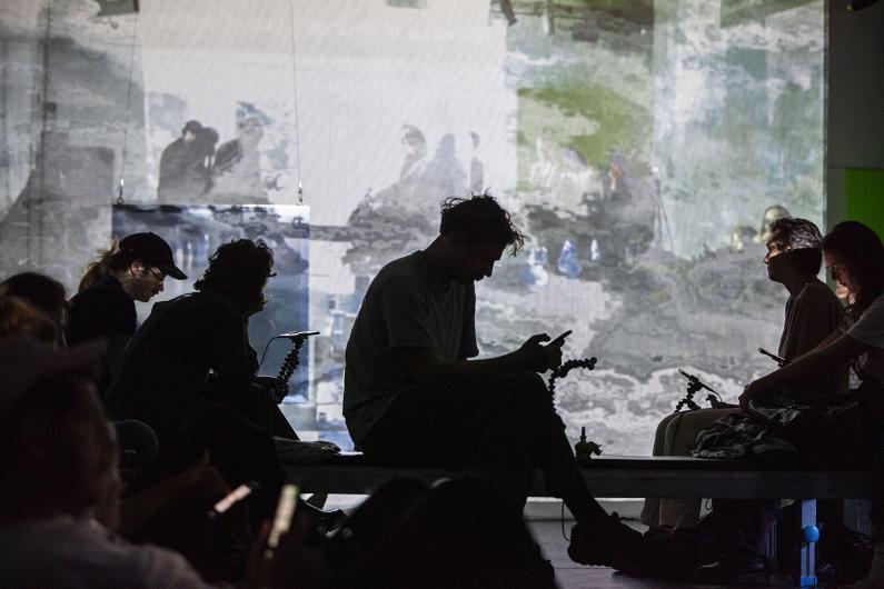 Silhouettes of people seated on benches in front of a screen displaying abstract, blurred visuals. The figures are partially illuminated by the soft glow of their phones and the projections, creating a contemplative and ambient scene.
