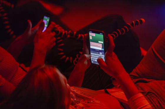 Nahaufnahme von Händen, die Smartphones halten, während Nachrichten auf einem grünen Bildschirm getippt werden. Die Szene ist in rotes Licht getaucht, und die Menschen scheinen sich in einer entspannten, informellen Umgebung zu befinden.