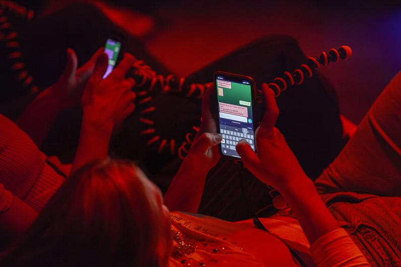 Close-up of hands holding smartphones, typing messages on a green screen. The scene is bathed in red light, and the individuals appear to be in a relaxed, informal setting.