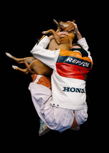A person in a striking Honda Repsol motorcycle jacket embraces a dog in an almost dancing pose. The background is completely black, making the scene almost surreal.