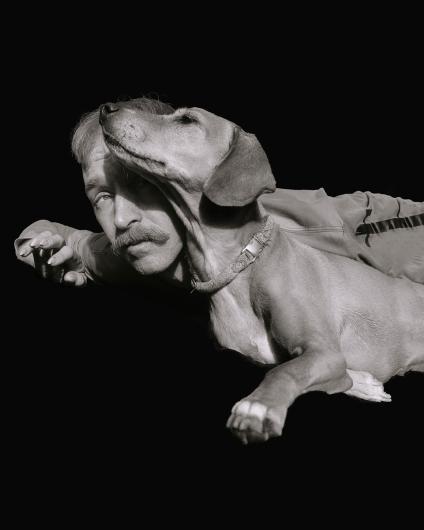 The head of a dog lies gently over the face of a person, revealing a tender connection between man and animal. The shot is in black and white and emphasizes the emotional depth of the moment.