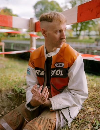 Sitting by a makeshift barrier, the person gazes pensively to the side. The motorcycle jacket is dirty, their hands are clasped — a snapshot that conveys both fragility and resilience.