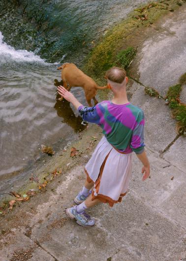 At the edge of a shallow stream, a person accompanies a dog exploring the water with curiosity. With an outstretched arm and open movements, a poetic image of trust, companionship, and playful ease emerges.