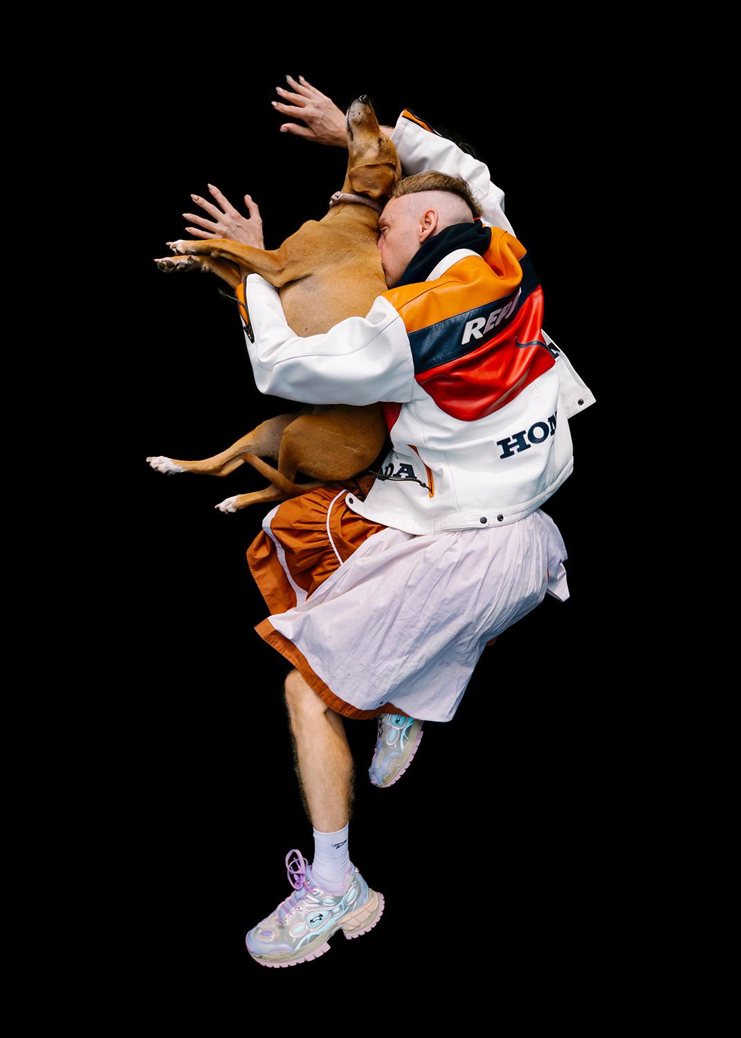 In einer kraftvollen, beinahe tänzerischen Bewegung hält die Person einen Hund eng umschlungen. Die farbenfrohe Repsol-Jacke, der schwebende Körper und der schwarze Hintergrund erschaffen ein surreal wirkendes Bild von Nähe und Freiheit.