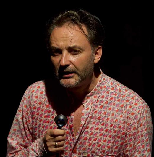 A man with short, dark hair and a beard is illuminated by a spotlight against a black background. He wears a patterned red and white shirt and holds a small round object, while speaking with an intense expression.
