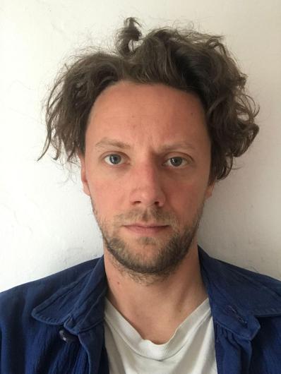 A man with messy, curly brown hair and blue eyes looks directly at the camera. He is wearing a white t-shirt under a blue jacket. The background is a plain white wall, and his expression is neutral.