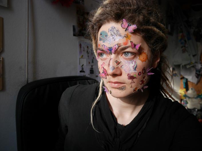 A person with wavy, light brown hair and blue eyes is sitting in a dimly lit room. Their face is covered with colorful stickers, including butterflies, animals, and other decorative elements. They have a serious expression and are dressed in black.