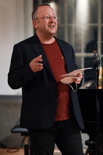 A middle-aged man with glasses, wearing a dark blazer over a red shirt, is speaking in front of an audience. He is holding a piece of paper and a face mask while gesturing with his hand. The setting appears to be an indoor event with a microphone visible.