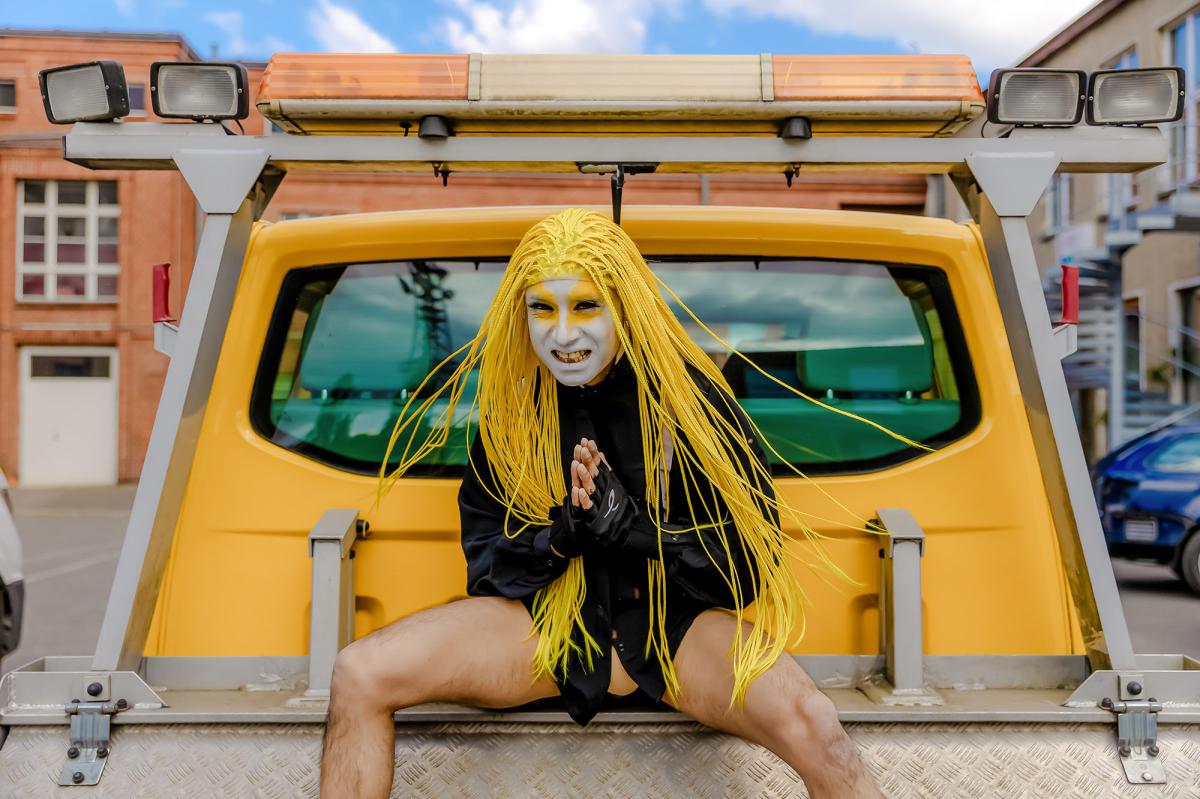 A person with long, bright yellow braided hair and white face paint with yellow eye makeup is sitting on the back of a yellow utility vehicle. They are wearing a black outfit and fingerless gloves. Their legs are spread apart, and they are holding their hands together in front of them with a dramatic expression, possibly grinning. The scene is set outdoors in an urban environment with buildings and cars in the background.