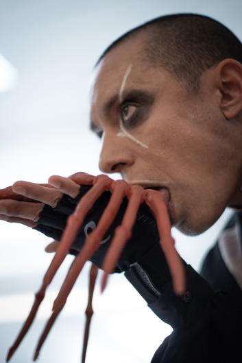 Eine Nahaufnahme einer Person mit auffälligem Bühnen-Make-up zeigt, wie er eine ungewöhnliche prothetische Hand mit verlängerten, tentakelartigen Fingern ableckt oder mit ihr interagiert. Das kontrastreiche Licht verstärkt die surreale, fast außerirdisch wirkende Ästhetik der Szene.