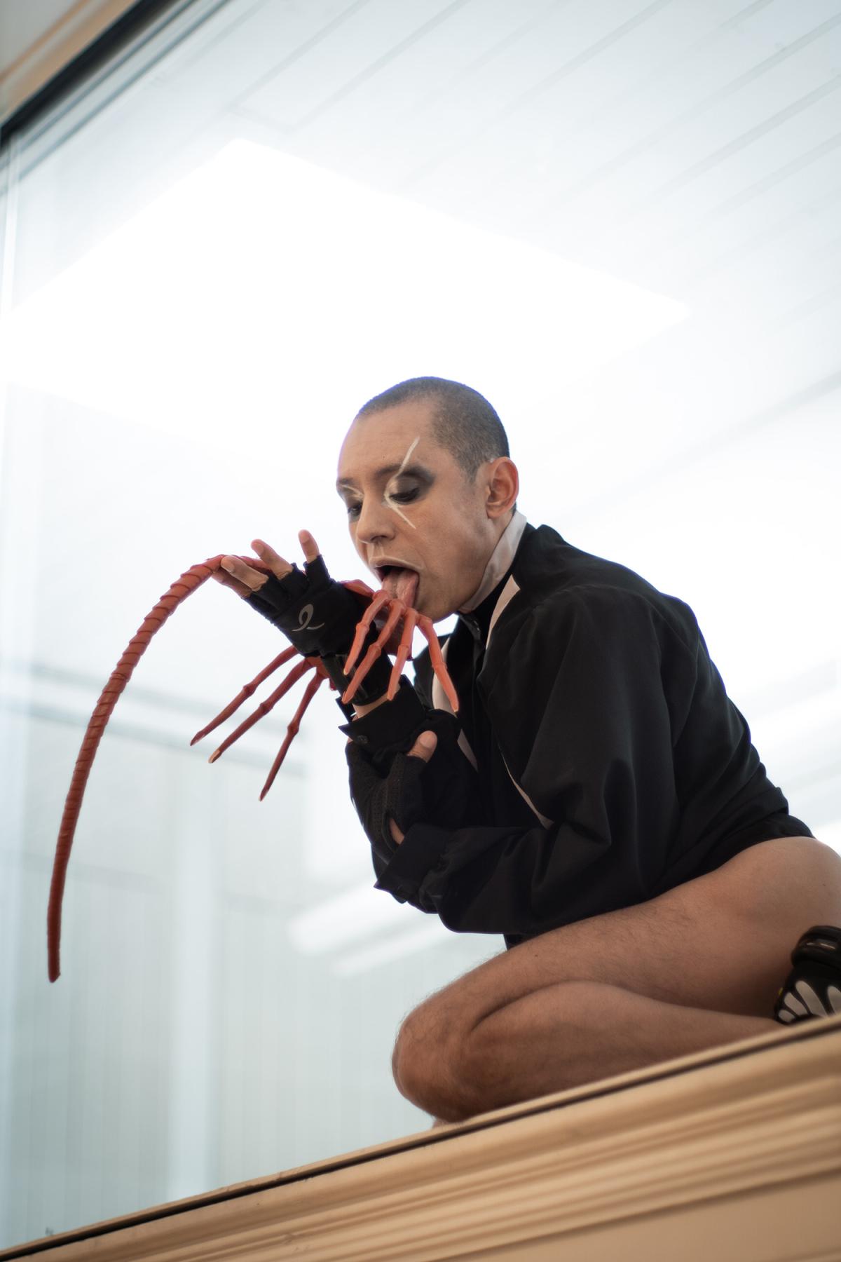 Eine Person mit theatralischem Make-up und rasiertem Kopf hockt auf einer Kante und leckt an einer verlängerten prothetischen Hand mit mehreren ausgestreckten Fingern. Er trägt ein schwarzes langärmeliges Hemd und fingerlose Handschuhe, was die unheimliche und avantgardistische Atmosphäre der Performance unterstreicht.