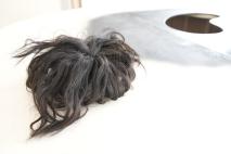 Ein Bündel dunkler, verfilzter Haare liegt auf einer weißen Oberfläche. Im Hintergrund befindet sich eine schwarze kreisförmige Form auf der Oberfläche, möglicherweise Teil einer Installation oder eines Kunstwerks. Die Umgebung wirkt minimalistisch mit einem starken Kontrast zwischen hellen und dunklen Elementen.