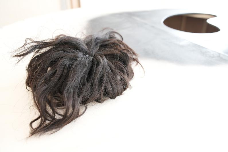 Ein Bündel dunkler, verfilzter Haare liegt auf einer weißen Oberfläche. Im Hintergrund befindet sich eine schwarze kreisförmige Form auf der Oberfläche, möglicherweise Teil einer Installation oder eines Kunstwerks. Die Umgebung wirkt minimalistisch mit einem starken Kontrast zwischen hellen und dunklen Elementen.