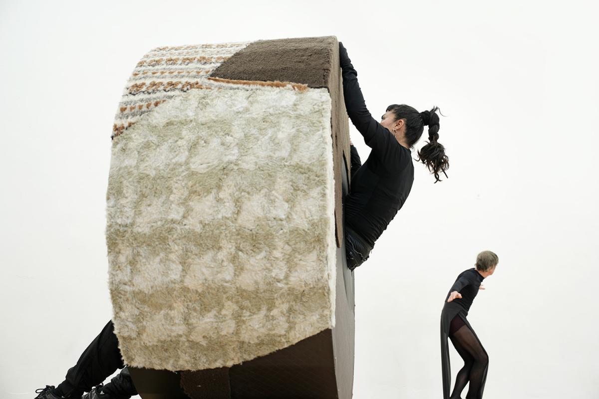 A performer dressed in black climbs a large, cylindrical structure covered with textured fabric, resembling a rolling object. Another person in the background, also in black clothing, moves dynamically. 