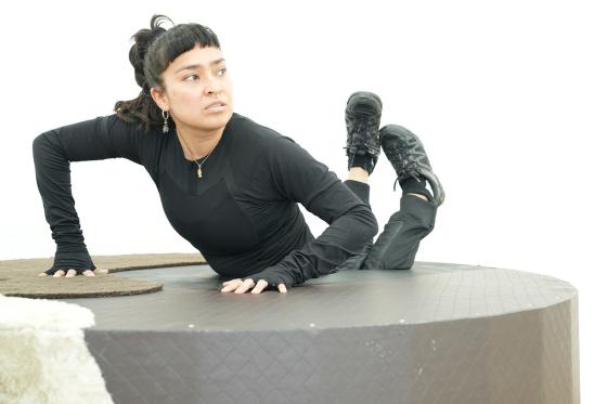 A performer dressed in black lies on top of a large, cylindrical structure covered in quilted fabric. Their expression appears focused, and they seem engaged in an act of balance or movement. The minimalist white background emphasizes the contrast with the dark clothing and the installation.