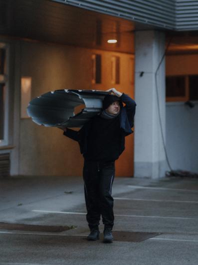 Eine Person in dunkler Kleidung trägt ein großes, gebogenes Metallstück auf der Schulter und geht durch einen Parkplatz im Freien.