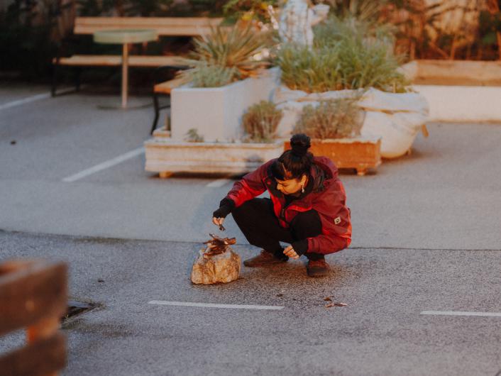 A person in a red jacket crouches in a parking lot, collecting small items from a rock.