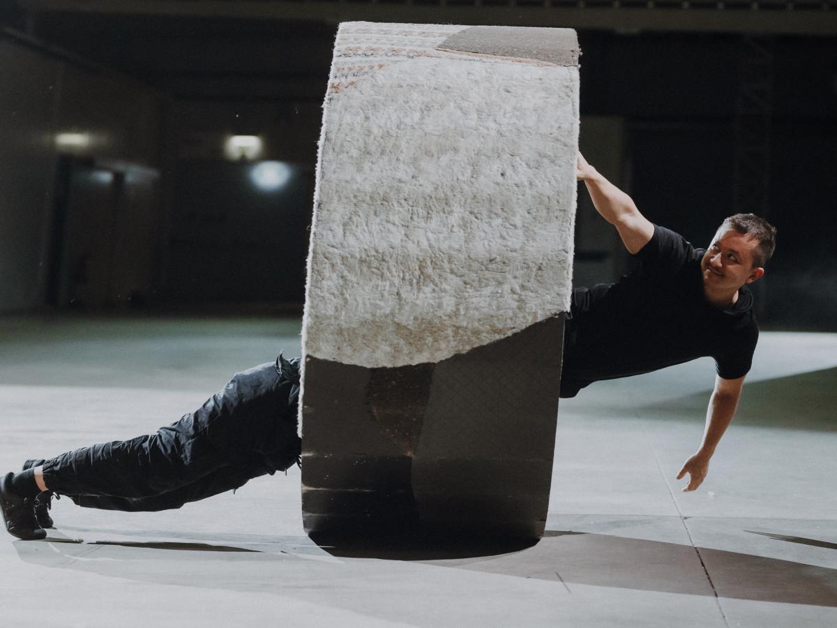 A person hangs sideways from a giant fabric-covered cylinder, while another person supports the wheel with their legs on the opposite side.