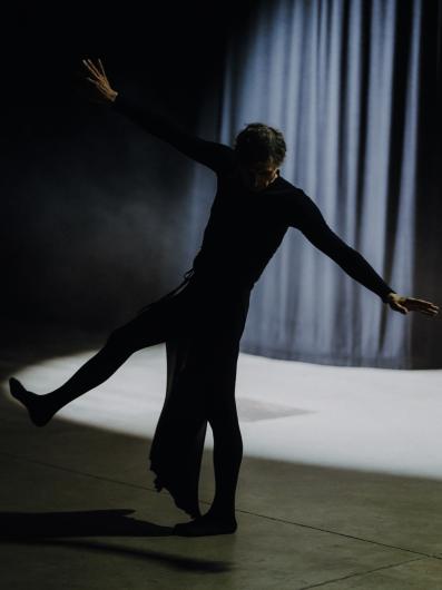 A person in black clothing dances alone on a stage, illuminated by a spotlight, with a dark background.