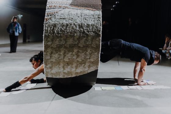 Zwei Personen ragen in einer Plank-Position beidseitig aus einem großen, mit Stoff bezogenen Zylinder und reichen nach Zetteln auf dem Boden.