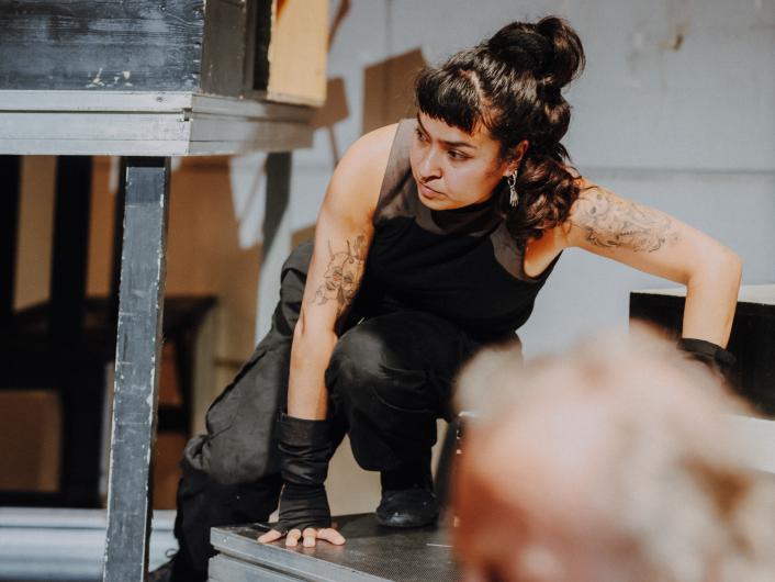 A person with tattooed arms and black gloves crouches on a stage structure, looking intently to the side.