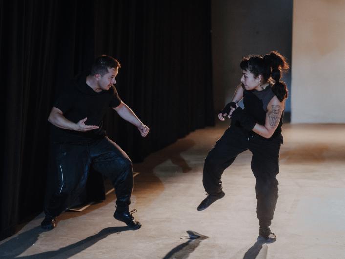 Two people dressed in black face each other on a stage in a mock fight scene, both in a combative stance.
