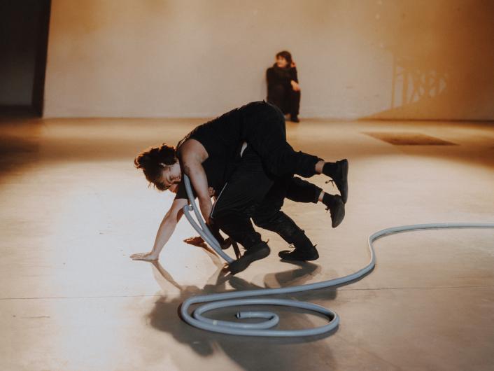 Two people become entangled in a dynamic movement with a long, flexible hose on the stage floor, while another person sits in the background.