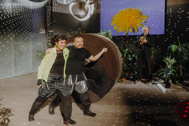 Two smiling people stand arm in arm in front of a large cylinder, while a person in the background plays the trumpet; the stage is decorated with plants and light effects.