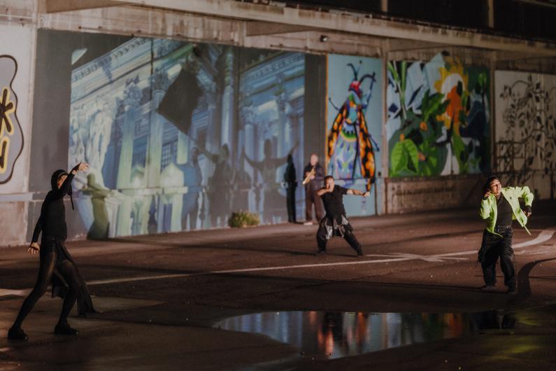Drei Personen führen auf einem nächtlichen Platz eine dynamische Performance vor bemalten Wänden und großen Projektionen durch; im Hintergrund sind bunte Wandbilder und ein Spiegelbild in einer Pfütze sichtbar.
