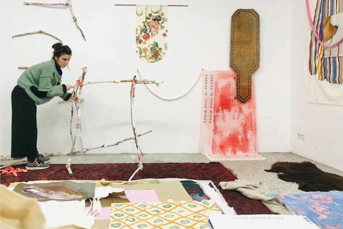 An artist wearing black trousers and a green knitted jumper works on a sculptural installation made of painted branches leaning against a white wall. Around her are rugs, fabrics, and art materials. Various artworks hang on the wall, including a floral textile piece, a banner reading “CHEESE IN YOUR WINGS,” and a woven object.