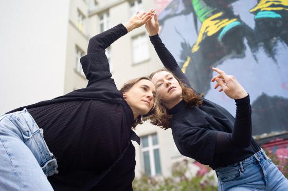 The photo shows the upper bodies of two white women in motion. The person who photographed them was standing a little lower down and took the picture from below. The women are wearing light-coloured jeans and black long-sleeved shirts. Their hair is loose and shoulder-length. They are both bent forwards. Their faces meet in the centre with a dreamy expression and gaze into the distance. One of the women's arms is organically placed above their heads and dances in front of an urban background of grey and colourful spray-painted house walls.