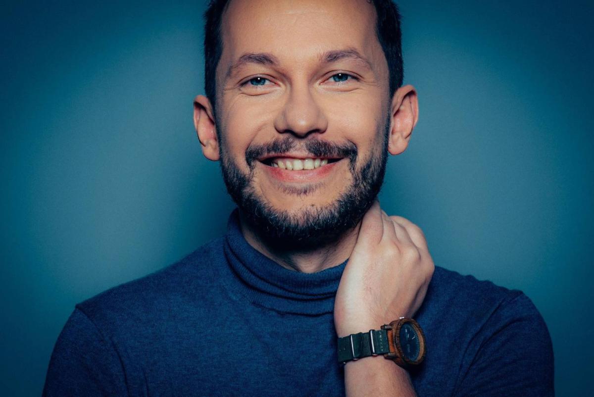 A smiling man looks directly at the camera. He has short dark hair, a well-groomed beard, and is wearing a dark blue turtleneck sweater. His right hand touches his neck, revealing a wristwatch with a wooden frame and dark face. The background is a solid dark blue. The lighting highlights his friendly face and creates a warm, welcoming atmosphere.