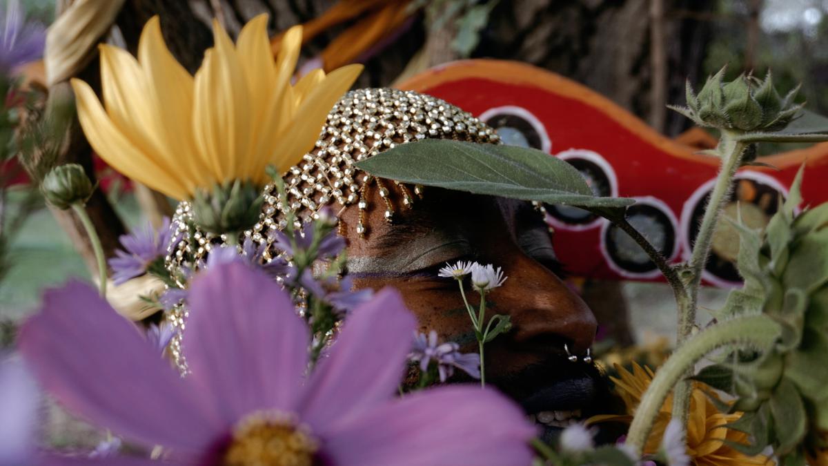 Close-up of a person whose face is partly obscured by flowers and leaves. Blurred purple blossoms, yellow petals and green plants fill the foreground. The person wears a glittering golden headpiece and striking make-up. In the background, a red decorative object with circular elements is visible. The composition feels dense, sensual and poetic, with a strong focus on textures, colours and floral details.