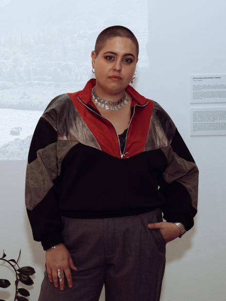 A person with very short hair stands in front of a white wall with exhibition texts. They wear a black, red, and grey jacket, a necklace with silver elements, and grey trousers. One hand is in their pocket, and they look calmly and directly into the camera.