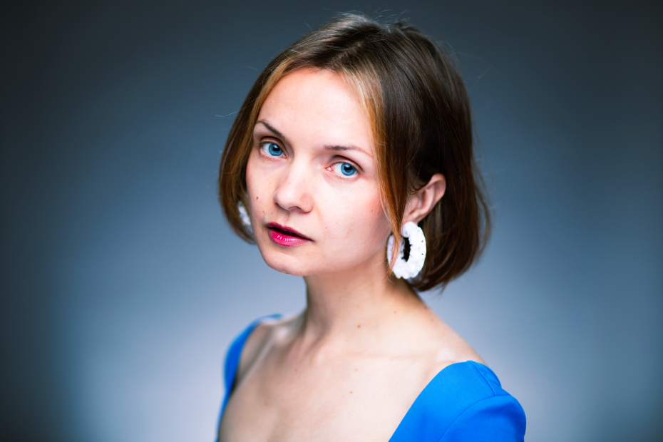 Close portrait of a person with short brown hair against a soft blue-grey background. They look directly into the camera and wear a bright blue top with a wide neckline, large white earrings and dark red lipstick. The light is soft and even, and the expression is calm and serious.