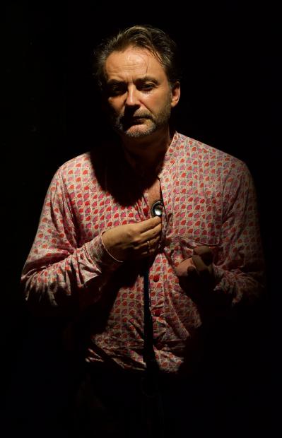 A person stands alone against a black background, lit from the front by a focused light. The person wears a patterned shirt in shades of pink and red and holds a stethoscope to their chest, as if listening to their heartbeat. Their expression appears serious and concentrated, with parts of the body fading into shadow. The atmosphere is quiet and intimate, with strong contrast between light and darkness.