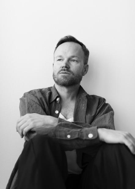Black-and-white portrait of a seated person against a plain light wall. The person wears an open shirt over a light top, with their arms loosely folded, gazing calmly slightly past the camera. The image feels quiet, focused and reflective.