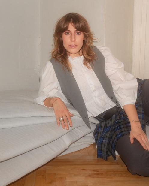A person with shoulder-length wavy hair leans casually against a wall on a stack of white mattresses, wearing a white ruffled blouse, gray vest, blue-and-black tartan skirt over dark trousers, with rings on their fingers and a small tattoo on their forearm; wooden floor and light-colored wall in the background.