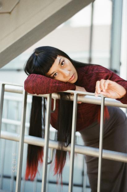 Portrait of Cat Jimenez wearing a fitted, mottled dark-red sweater and high-waisted dark trousers, leaning casually with one arm on a light grey metal railing; their long, straight black hair with reddish ends hangs down through the bars, a blunt fringe framing their face, which is tilted slightly toward the camera with a serious, thoughtful expression; in the background, windows and architecture blur into soft daylight.