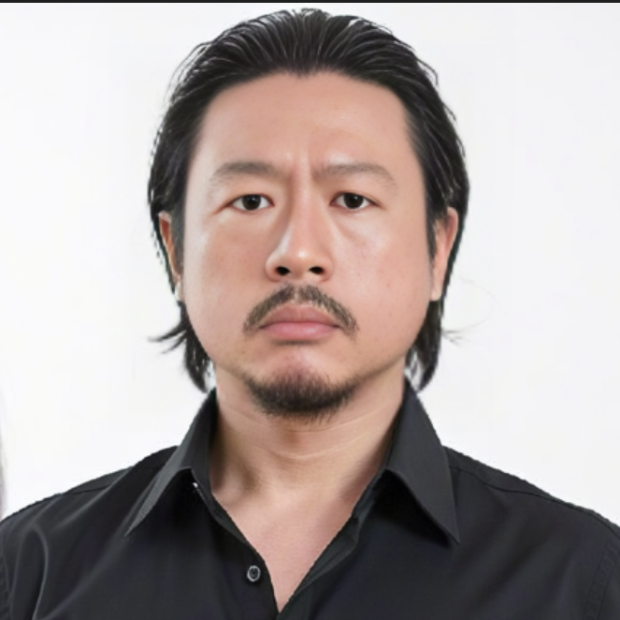 Portrait of a person against a light, neutral background. The person looks directly into the camera and wears a black shirt with an open collar. Their dark, smooth hair is brushed back. A narrow moustache and a short beard on the chin frame the face. The expression appears calm and serious.