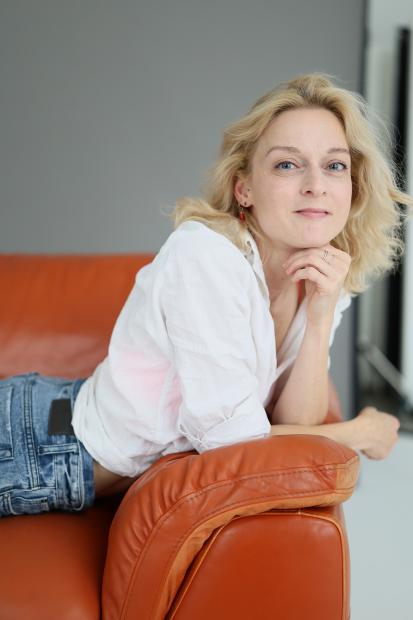 Eine Person lehnt seitlich auf einer braunen Ledercouch und blickt ruhig in die Kamera. Der Kopf ist auf eine Hand gestützt, der Körper liegt entspannt auf der Couch. Die Person trägt ein helles Oberteil und Jeans. Der Hintergrund ist schlicht und hell, wodurch die Person im Vordergrund klar hervorgehoben wird. Weiches Tageslicht sorgt für eine ruhige, intime Atmosphäre.