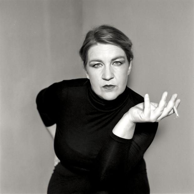 Black-and-white portrait of a person in a black turtleneck against a plain background. The person leans forward, looks directly into the camera and holds a cigarette in their outstretched hand.
