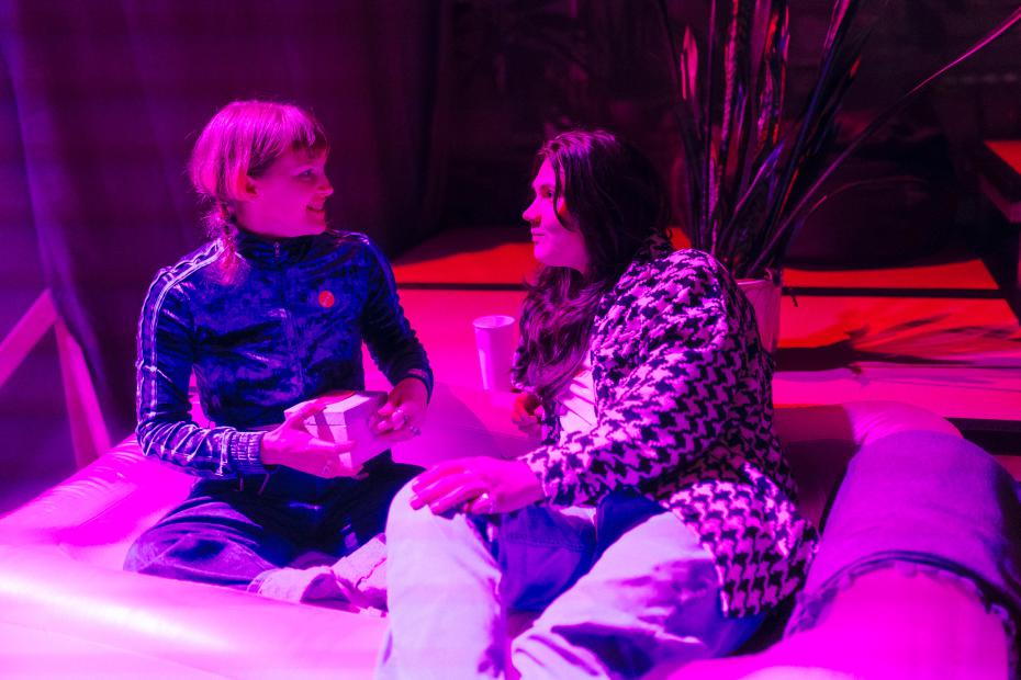Two people sit facing each other on a sofa in a room lit with pink and purple light. They look at one another, appearing engaged in conversation. One person wears a blue zip-up tracksuit and holds a small light-coloured box in their hands. The other person wears a patterned jacket and jeans and leans slightly forward. Plants and a cup are visible in the background, creating an intimate, relaxed and domestic atmosphere.