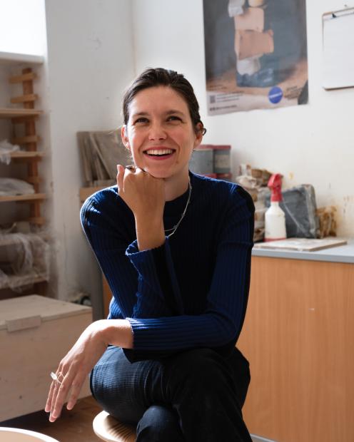 A person sits on a stool in a bright workspace and smiles toward the camera. Their head rests on one hand, with the upper body leaning slightly forward. The person is wearing a dark blue top and dark trousers. Shelves, work materials, and a work surface in the background suggest a studio or workshop setting. Daylight enters the room from the side.