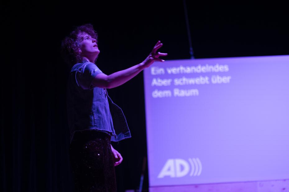 F emerges from the darkness into pink-purple light. The head slightly raised, the gaze as is awe, one hand stretching out in front. Behind F, the abbreviation “AD” for audio description is projected onto a purple screen, along with the slightly blurred German words: “ Ein verhandelndes Aber schwebt über dem Raum.”
