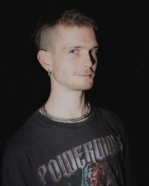 Porträt einer Person mit sehr kurzem Haar vor dunklem Hintergrund. Sie trägt ein dunkles T-Shirt mit Aufdruck, eine silberne Kette und Ohrringe und blickt ruhig zur Kamera.