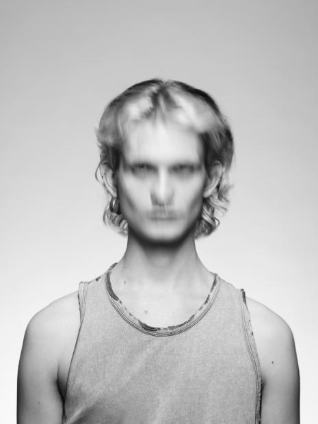 Schwarz-weiß-Porträt einer Person mit schulterlangem, leicht welligem Haar, die frontal in die Kamera blickt. Gesicht und Hals sind durch Bewegungsunschärfe stark verwischt, die Augen nur schemenhaft erkennbar, was der Aufnahme einen geisterhaften, experimentellen Charakter gibt. Die Person trägt ein ärmelloses, leicht abgenutzt wirkendes Tanktop mit ausgefranstem Halsausschnitt und sichtbaren Nähten vor einem neutralen, hellen Hintergrund.